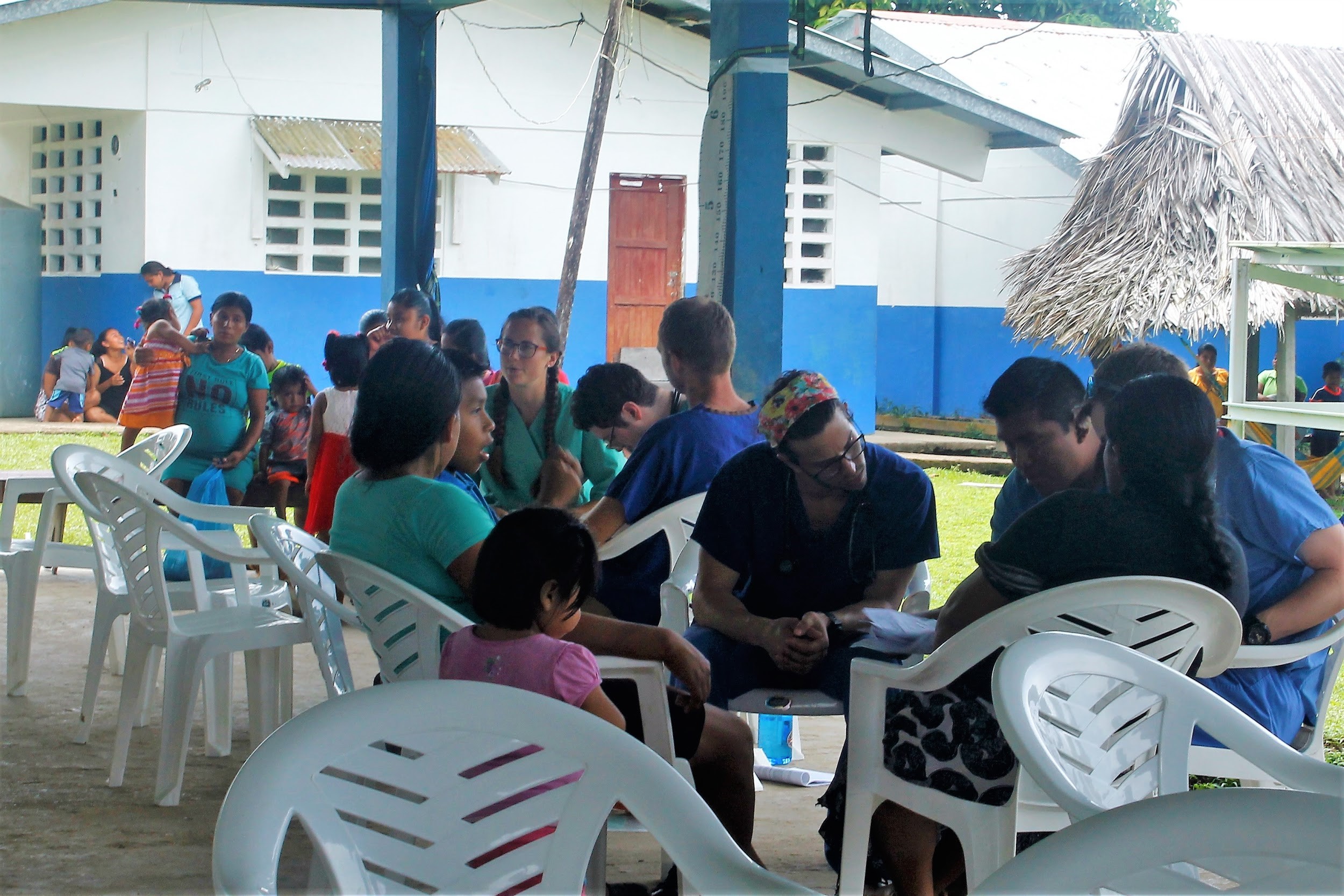 First-year medical student Corey Kronman, center, wearing a headband, talks to patients at a mobile clinic in Panama. He is looking at a paper with a man. Behind him is a plastic table with a woman medical student talking to her patients. In the background a group of indigenous Ngabe people stand or sit as they wait. A thatched hut is on the right, and a cement block building is behind them.