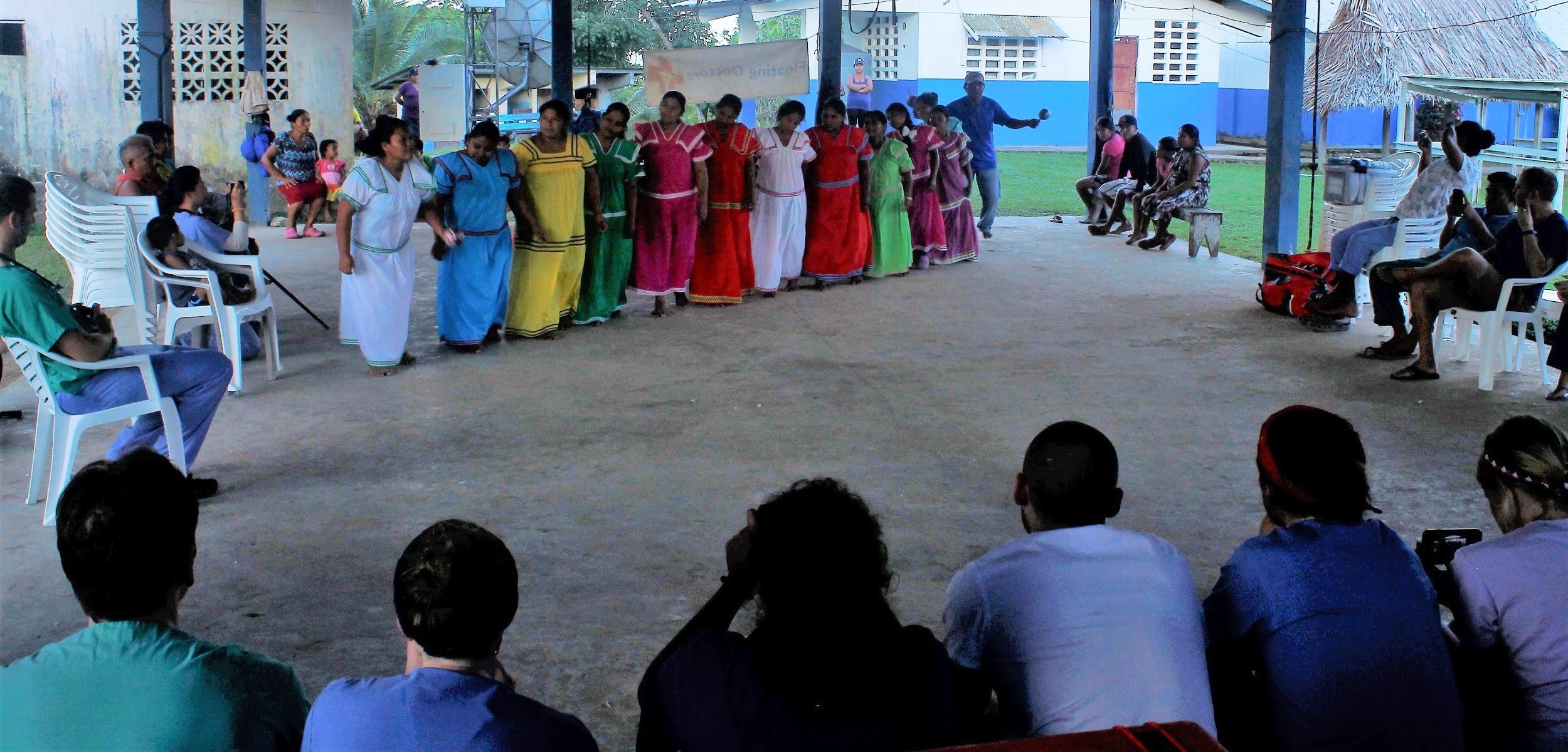 A group of 11 women of the Ngabe community stand in a row under a pavilion. They are wearing colorful dresses and performing a dance of appreciation for the Penn State College of Medicine students. Students and natives sit in the foreground watching. Two rows of people sit on either side watching.