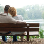 A man and a woman sit side by side on a wooden bench, facing away from the camera. The man’s arm is around the woman. They look out at a lake surrounded by trees.