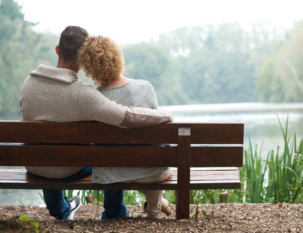 A man and a woman sit side by side on a wooden bench, facing away from the camera. The man’s arm is around the woman. They look out at a lake surrounded by trees.