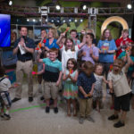 Several people -- more than a dozen children and two adults -- share space on a TV set. The Children's Miracle Network logo is in the background on the right; a TV monitor with the figure "$4,445,580" is on the left.