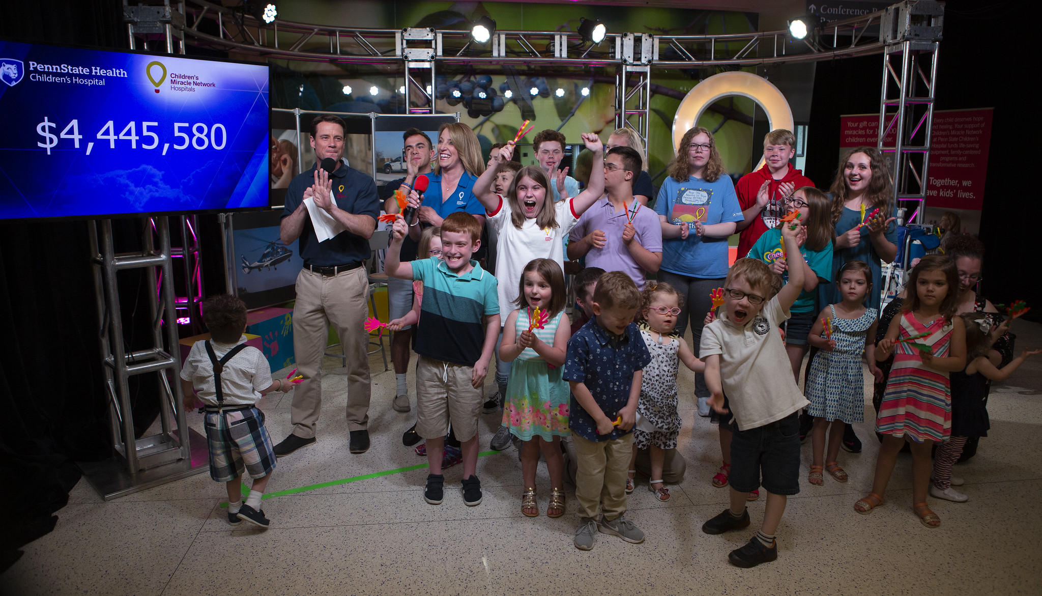 Several people -- more than a dozen children and two adults -- share space on a TV set. The Children's Miracle Network logo is in the background on the right; a TV monitor with the figure 