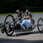 Cody Wills, wearing a helmet and sunglasses, rides his hand cycle, lying on his back, his fists grip handles that that push the bike forward on three wheels.