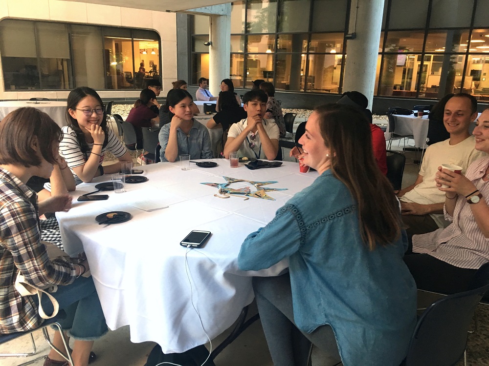 Eight international students with the 2018 Global Health Exchange Program sit at a round table in a restaurant inside Penn State College of Medicine. They are in their 20s, smiling and wearing casual clothes. Behind them people sit at other tables. Large columns and a bank of windows are in the background.