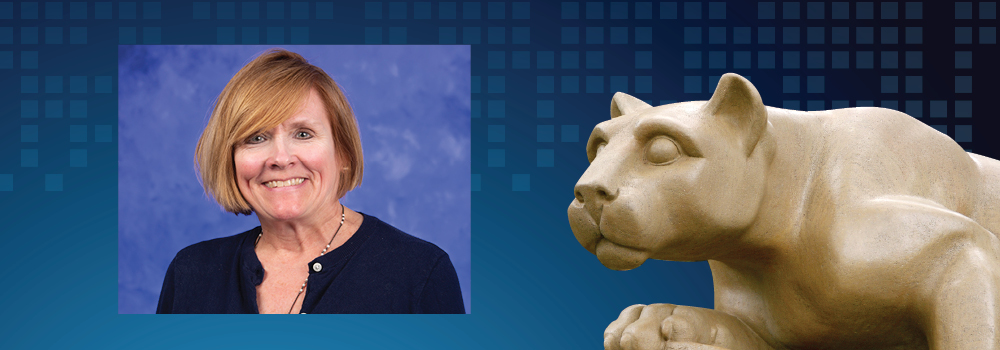 Judy Hlafcsak, wearing a dark top and a necklace, smiles. A statue of the Penn State Nittany Lion appears to her right.