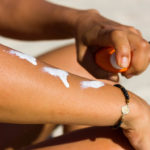 Close-up view of a woman’s right arm, as her left hand uses a quirt bottle to apply white sunscreen to the right arm. Sand is in the background, out of focus.