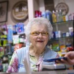 Arlene Faul, a 95-year-old woman from Hershey, smiles behind the counter of the Hershey Medical Center gift shop. She is wearing a vest, shirt, necklace and glasses. A man’s hand holds a phone out to her. Behind her are shelves of toiletries and other items. A top and sweater hang on a hanger on the right.
