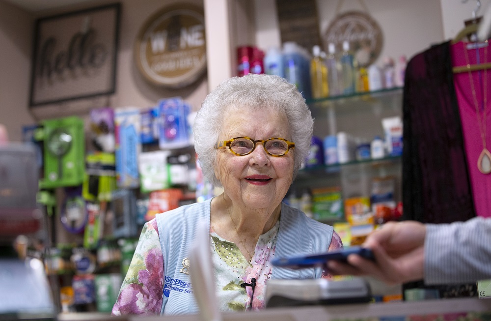 Arlene Faul, a 95-year-old woman from Hershey, smiles behind the counter of the Hershey Medical Center gift shop. She is wearing a vest, shirt, necklace and glasses. A man’s hand holds a phone out to her. Behind her are shelves of toiletries and other items. A top and sweater hang on a hanger on the right.