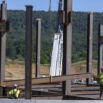 Two men in hardhats position a steel beam at a construction site. Behind them is broken ground. Tree-covered mountains rise into a clear sky.