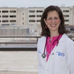 Dr. Karen Krok stands on the roof of Penn State College of Medicine smiling and wearing a white lab coat and stethoscope around her neck. The Hershey Medical Center logo is on the right pocket of her lab coat. She has medium-length brown hair. Behind her a building is under construction.
