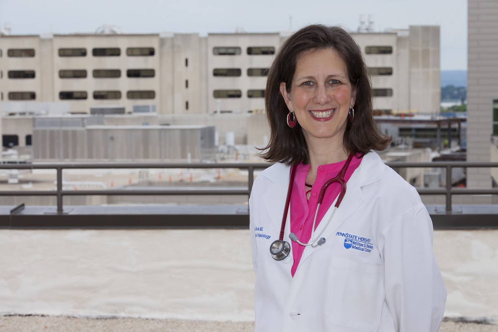 Dr. Karen Krok stands on the roof of Penn State College of Medicine smiling and wearing a white lab coat and stethoscope around her neck. The Hershey Medical Center logo is on the right pocket of her lab coat. She has medium-length brown hair. Behind her a building is under construction.