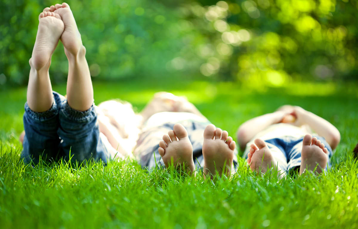 Three young people lay side by side in a grassy area. The photo is taken from the ground, viewing their feet. The person on the left has their feet raised into the air. A line of trees is in the background, out of focus.