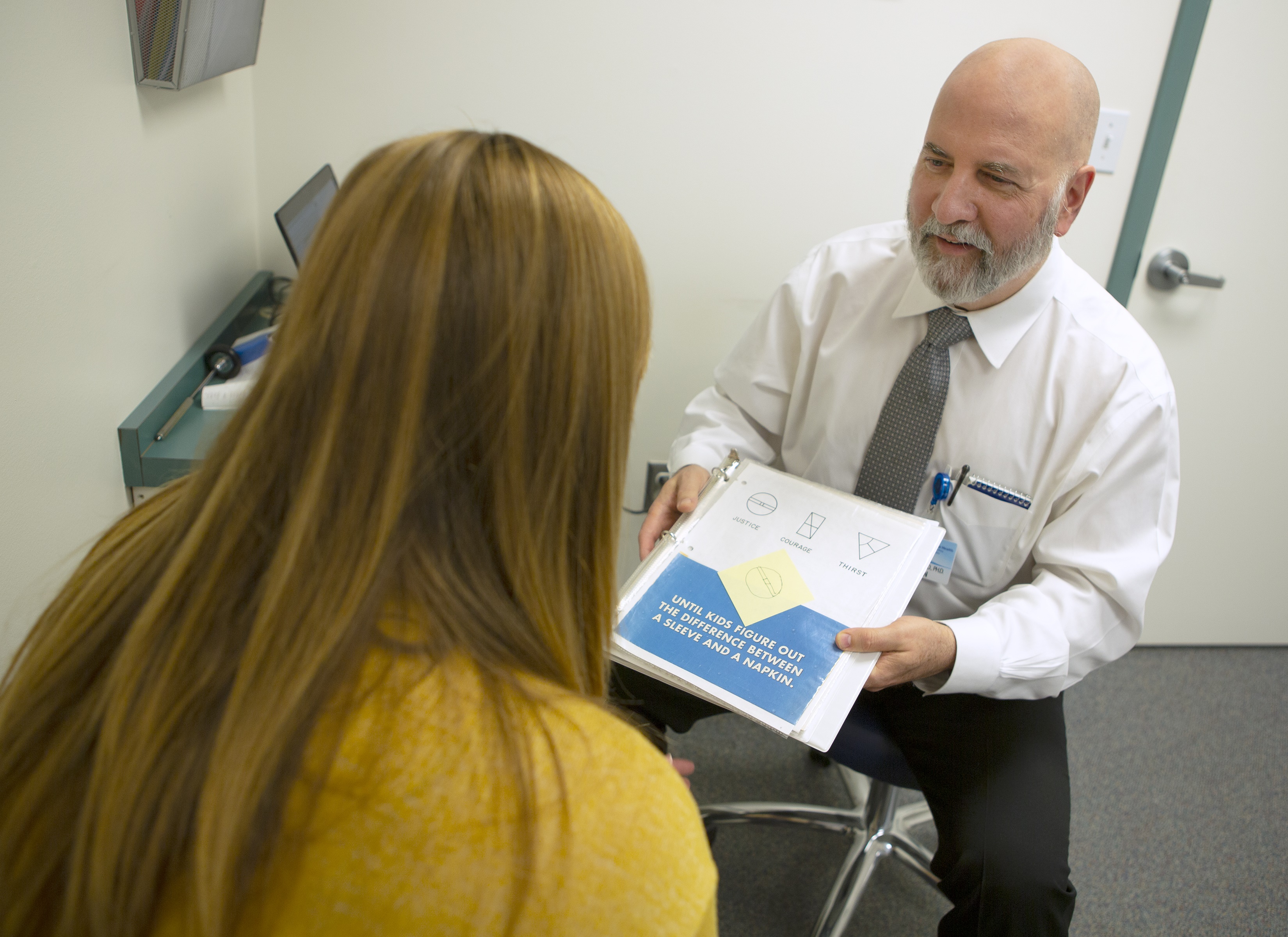 A male physician wearing a tie sits on a stool in an exam room, displaying a page with various diagrams on it to a female patient who sits across from him.