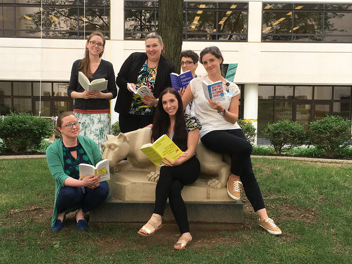 A group of six women are pictured sitting on and standing near a statue of the Penn State Nittany Lion in an outdoor courtyard. Each woman is holding a book.