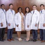 Six men and women in lab coats smile at the camera as they stand in front of a wood wall.