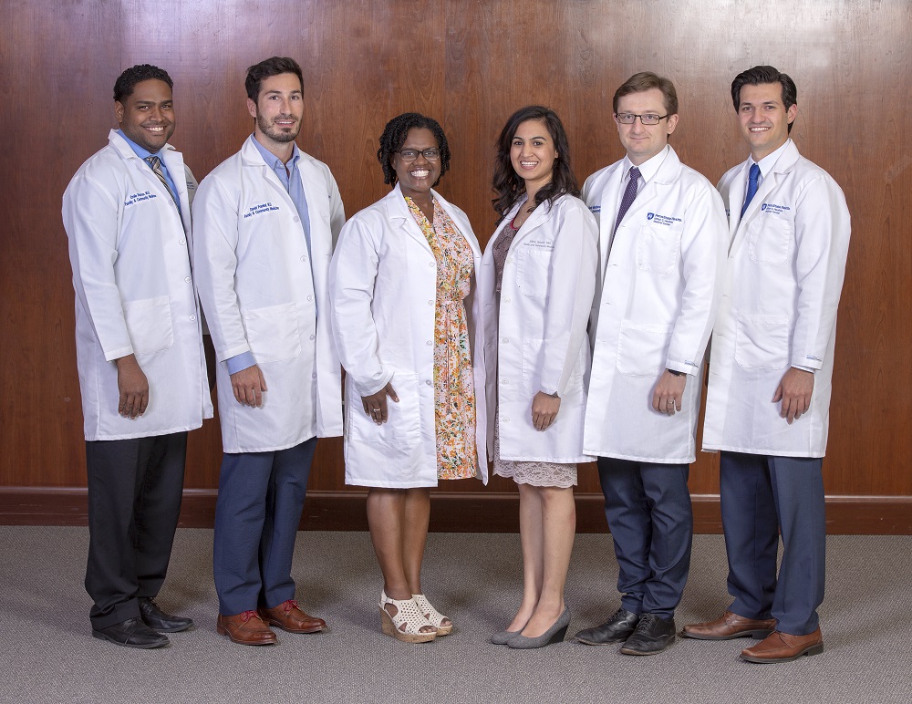 Six men and women in lab coats smile at the camera as they stand in front of a wood wall.