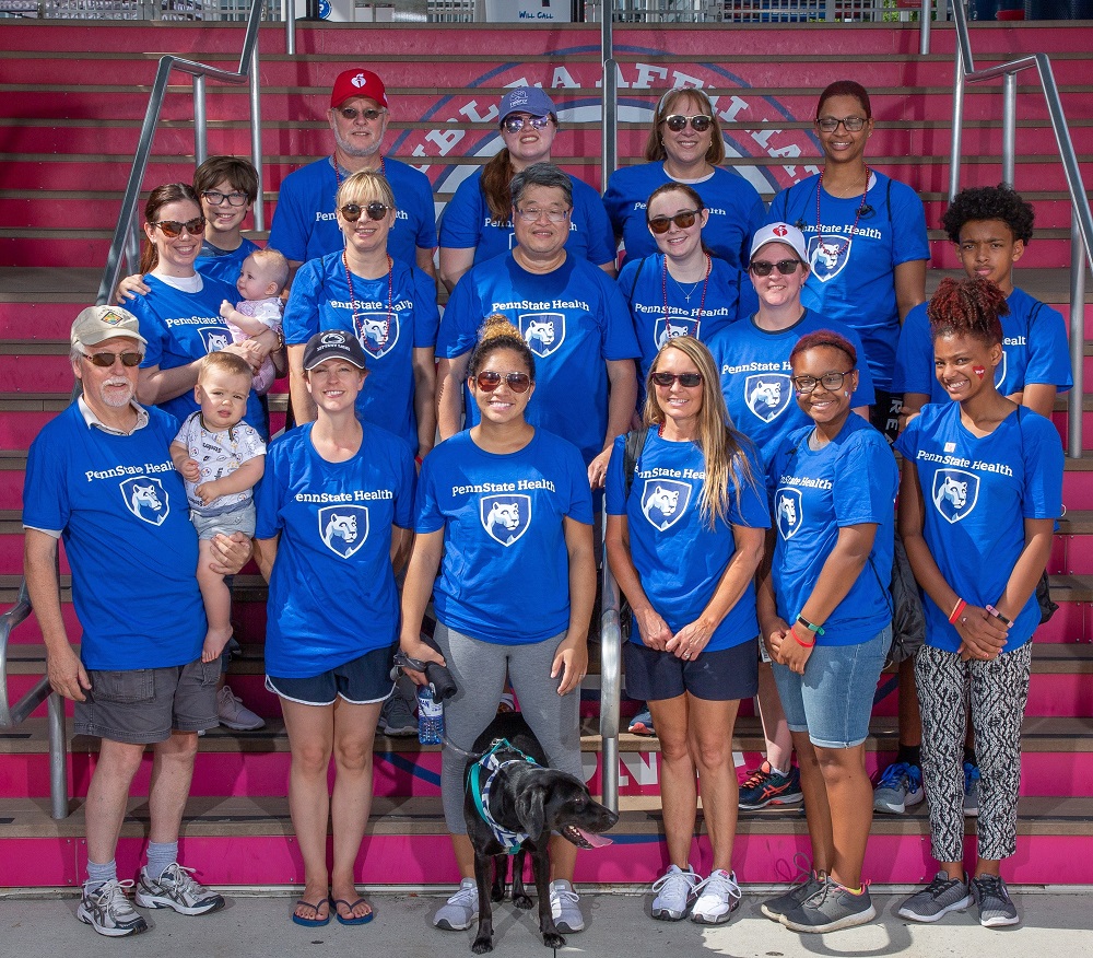 A group of 19 people, including Penn State Health employees and their family members, pose at the 2018 Heart Walk. They are wearing Penn State Health T-shirts and standing on stairs. Two babies and a Black Labrador Retriever are with them.