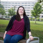 Alissa Meister sits on a bench on the lawn of Penn State College of Medicine, which is seen in the background.