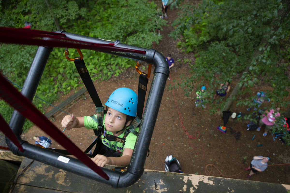 View from the top of a rock wall, looking down, as a young girl in climbing gear and supported by a square metal frame climbs the wall. About 6 people are on the ground, looking up.