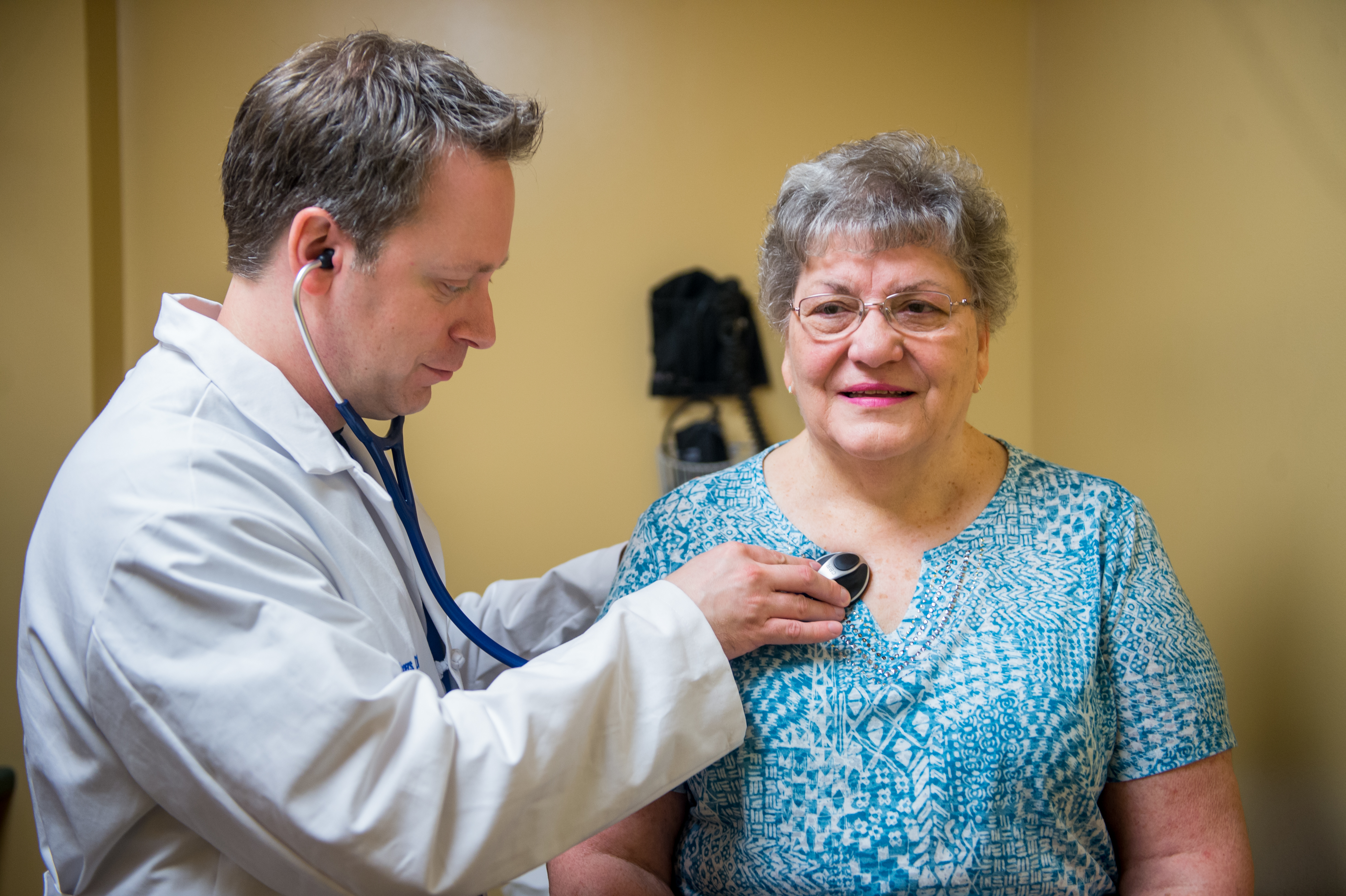 A male physician in a white coat uses a stethoscope to examine a female patient. Small medical equipment is mounted to a wall in the background.
