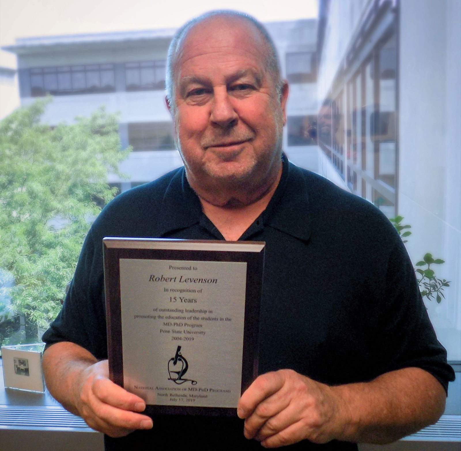 Dr. Robert Levenson of Penn State College of Medicine is seen in front of a window, holding a plaque.
