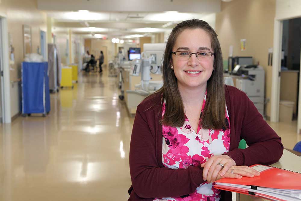 Mary Codi, a medical social worker at Penn State Health St. Joseph, stands in a hallway at St. Joseph Medical Center. She is leaning on a table and has an orange folder in front of her. She is wearing glasses, a sweater and a flowered top. She has medium-length straight hair. Behind her hospital equipment and rooms are visible.