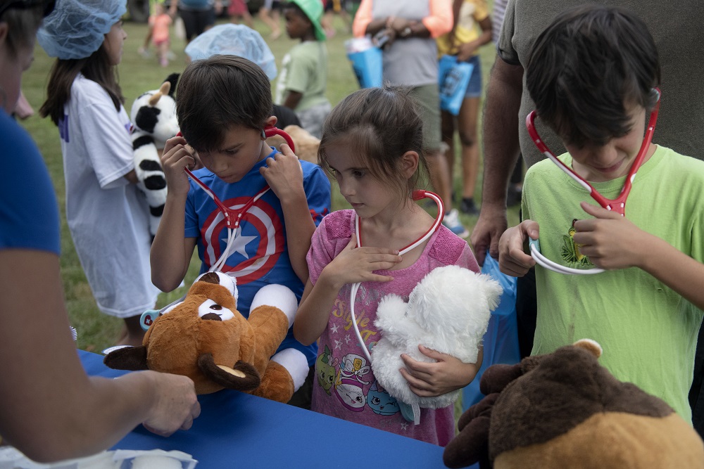 Three children stand in a row and wear stethoscopes. Two of the children hold stuffed animals .