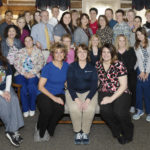 A room full of people pose for a photo. Some sit in chairs. Others stand.