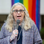 Rachel Levine speaks into a microphone. Behind her, a rainbow-colored flag is draped.