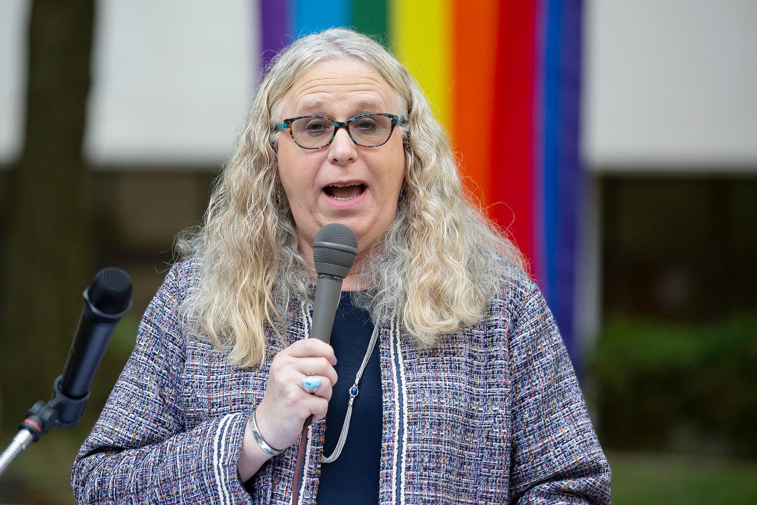 Rachel Levine speaks into a microphone. Behind her, a rainbow-colored flag is draped.