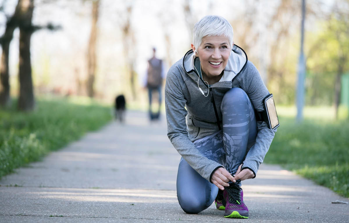 A woman in running tights and a light jacket kneels to tie her left shoe. She is on a paved walking path bordered by grass, and a person and dog are in the background, out of focus. A smart phone is strapped to the woman’s left arm, and she has ear buds in.
