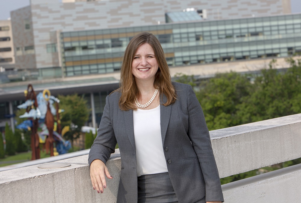 Deborah Berini, Hershey Medical Center president, leans on a wall overlooking the medical center. Behind her is a large metal sculpture. She is wearing a suit and a pearl necklace.