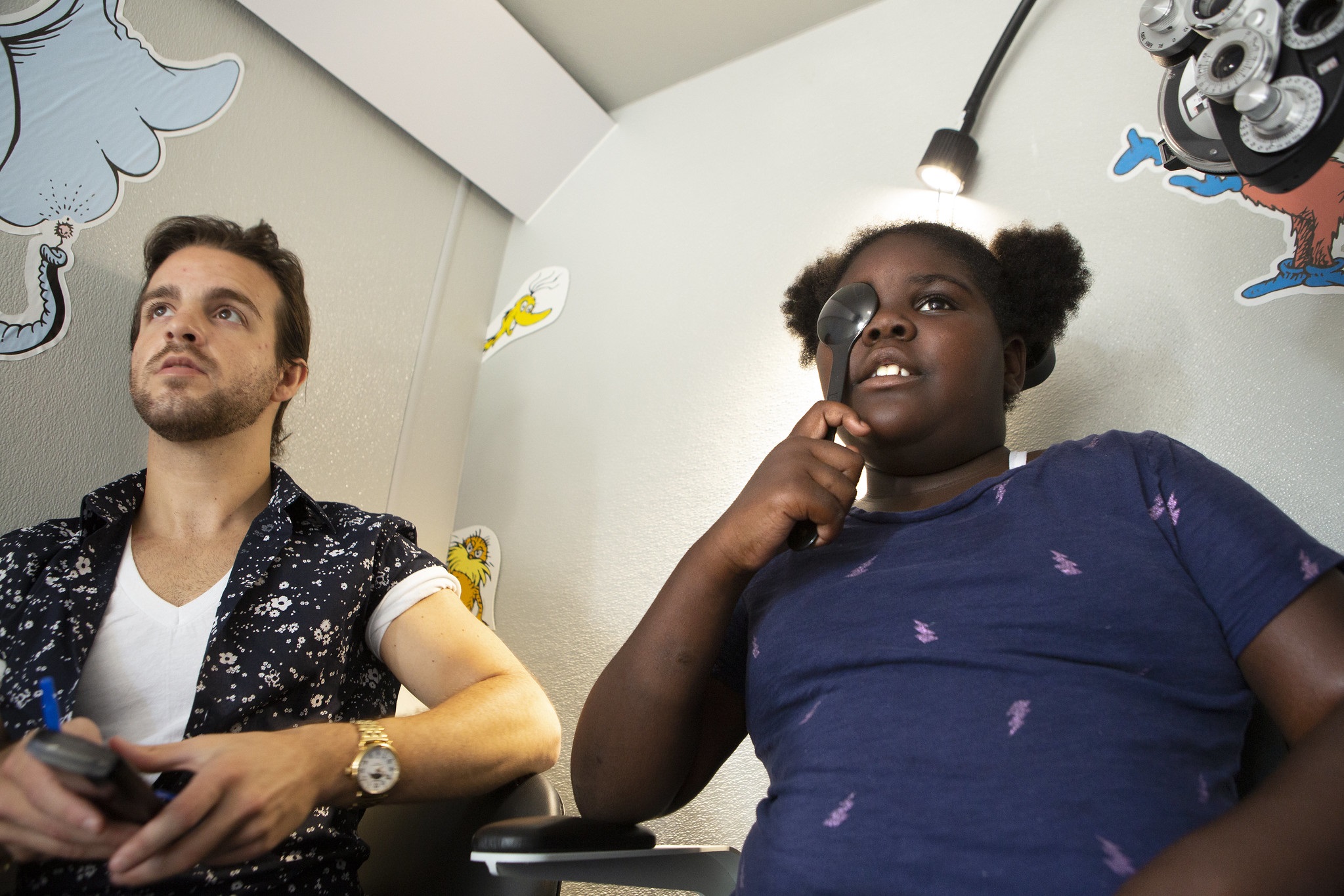 Dr. Aaron Harsch, an optometrist with Vision to Learn’s mobile eye clinic, sits next to 89-year-old Paris Gentry as she holds an eye occluder to her eye. She has curly hair and is wearing a short-sleeved top. Behind them Dr. Seuss characters are on the wall. Eye testing equipment is to the upper-right.
