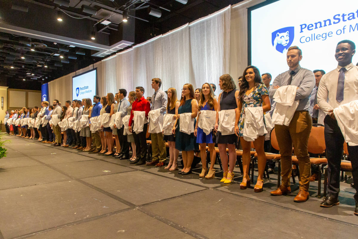 Several dozen medical students stand in two rows across a stage, with white medical coats draped over their left arms. The Penn State College of Medicine logo is projected on two screens behind them, and just in front of a pipe-and-drape backdrop.