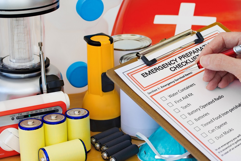 A handholds a pen to a checklist on a clipboard. The checklist is entitled “Emergency Preparedness Checklist.” Behind the clipboard, spread out an a table are a lantern, a flashlight, a radio, batteries, a first-aid kit and canned goods.