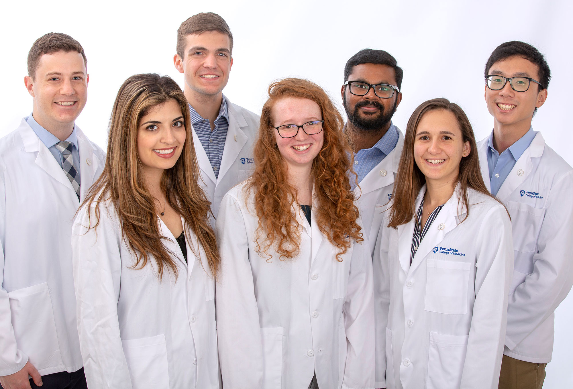A group of seven college students are seen wearing white medical coats against a professional photo backdrop.