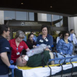 Several people in medical attire stand alongside a gurney on which a man lays down with his hands crossed over him. The word "actor" is on a tag hanging near his face. The Hershey Medical Center building and several other medical personnel are in the background.