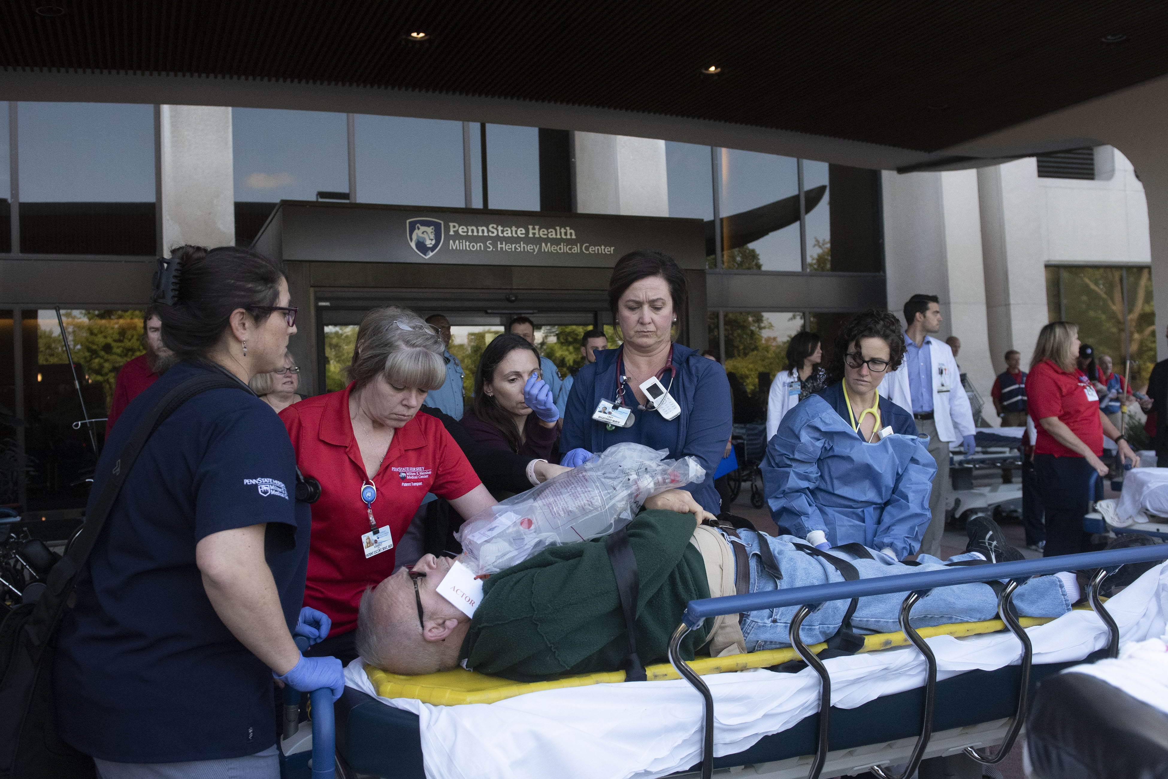 Several people in medical attire stand alongside a gurney on which a man lays down with his hands crossed over him. The word 