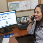 Belkys Perez talks into a telephone receiver while seated at a desk. A monitor next to her holds an image of the Penn State Health infonet. A lap top next it shows a tree-lined street. Behind both screen is a bulletin board posted with maps and other paper.