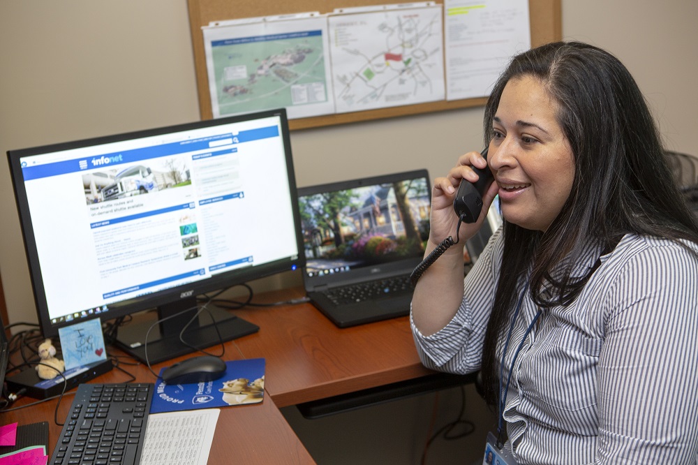 Belkys Perez talks into a telephone receiver while seated at a desk. A monitor next to her holds an image of the Penn State Health infonet. A lap top next it shows a tree-lined street. Behind both screen is a bulletin board posted with maps and other paper.