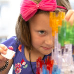 Close-up of a young girl with a bow in her hair building a multi-colored marble tunnel out of plastic building blocks.