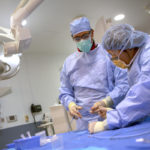 Two medical personnel wearing surgical scrubs, masks and caps work in an operating room. In the background is an overhead light and a clock on the wall, along with other equipment.