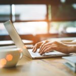 A man’s hands type on a laptop which is sitting on a desk. The man is wearing a wristwatch. A bonsai tree and a coffee cup are on the desk. Sunspots are seen on the coffee cup.