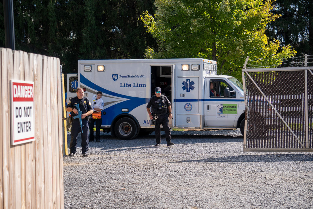 Two officers armed with "exercise" weapons stand just outside of an open gate and in front of a Penn State Health Life Lion ambulance. A Life Lion staff member stands alongside the vehicle.