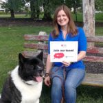 Deborah Berini sits on a bench holding her dog Alvin’s leash and a sign that says “Join our team!” She is wearing a polo shirt and jeans and has shoulder-length hair. Her dog is large and has black and white fur. Behind them are trees and grass.
