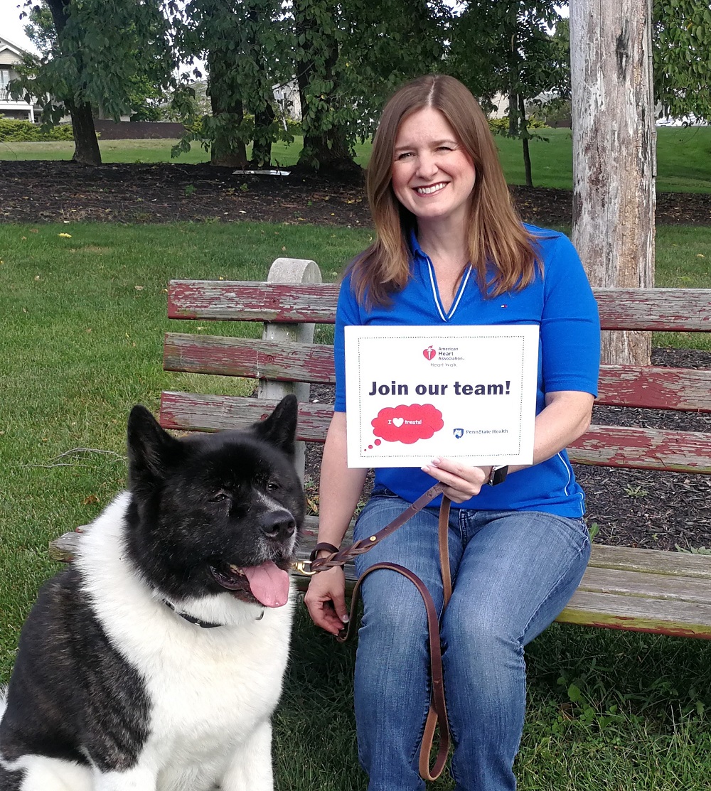 Deborah Berini sits on a bench holding her dog Alvin’s leash and a sign that says “Join our team!” She is wearing a polo shirt and jeans and has shoulder-length hair. Her dog is large and has black and white fur. Behind them are trees and grass.