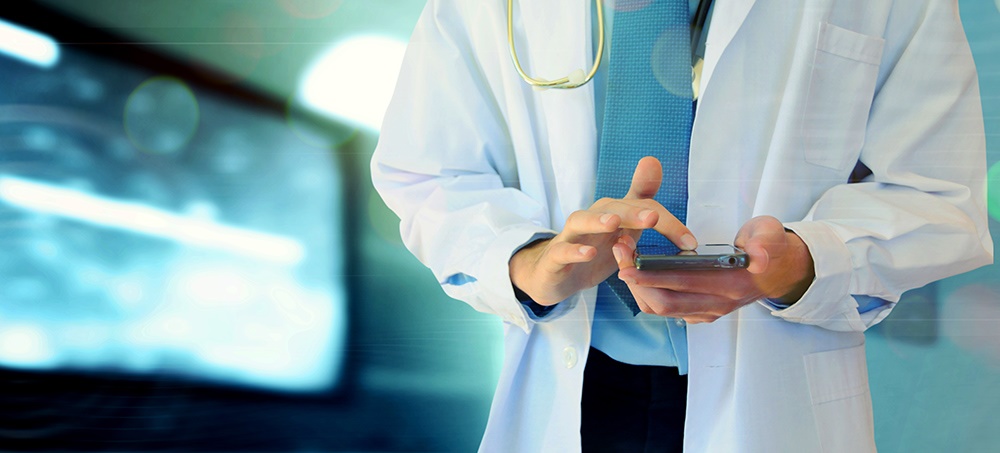 A male physician in a white lab coat touches the screen of a smartphone. He has a stethoscope over his shoulder. Behind him on the left is a monitor with an abstract image on it.