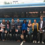 Thirteen people dressed casually stand in a row in front of a bus that has a sign “The Pennsylvania State University” on it. A male professor on the left is wearing a lanyard and name badge.