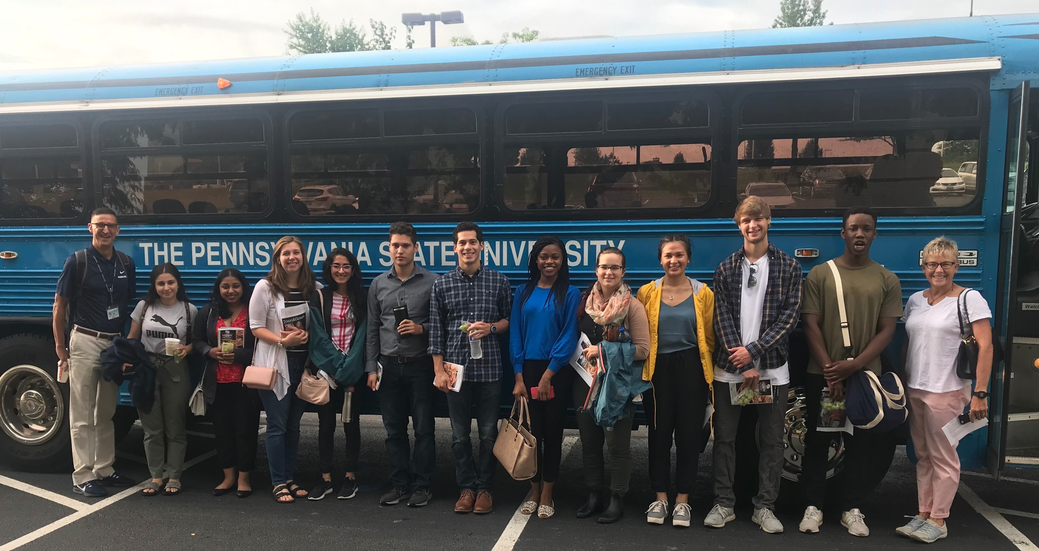 Thirteen people dressed casually stand in a row in front of a bus that has a sign “The Pennsylvania State University” on it. A male professor on the left is wearing a lanyard and name badge.