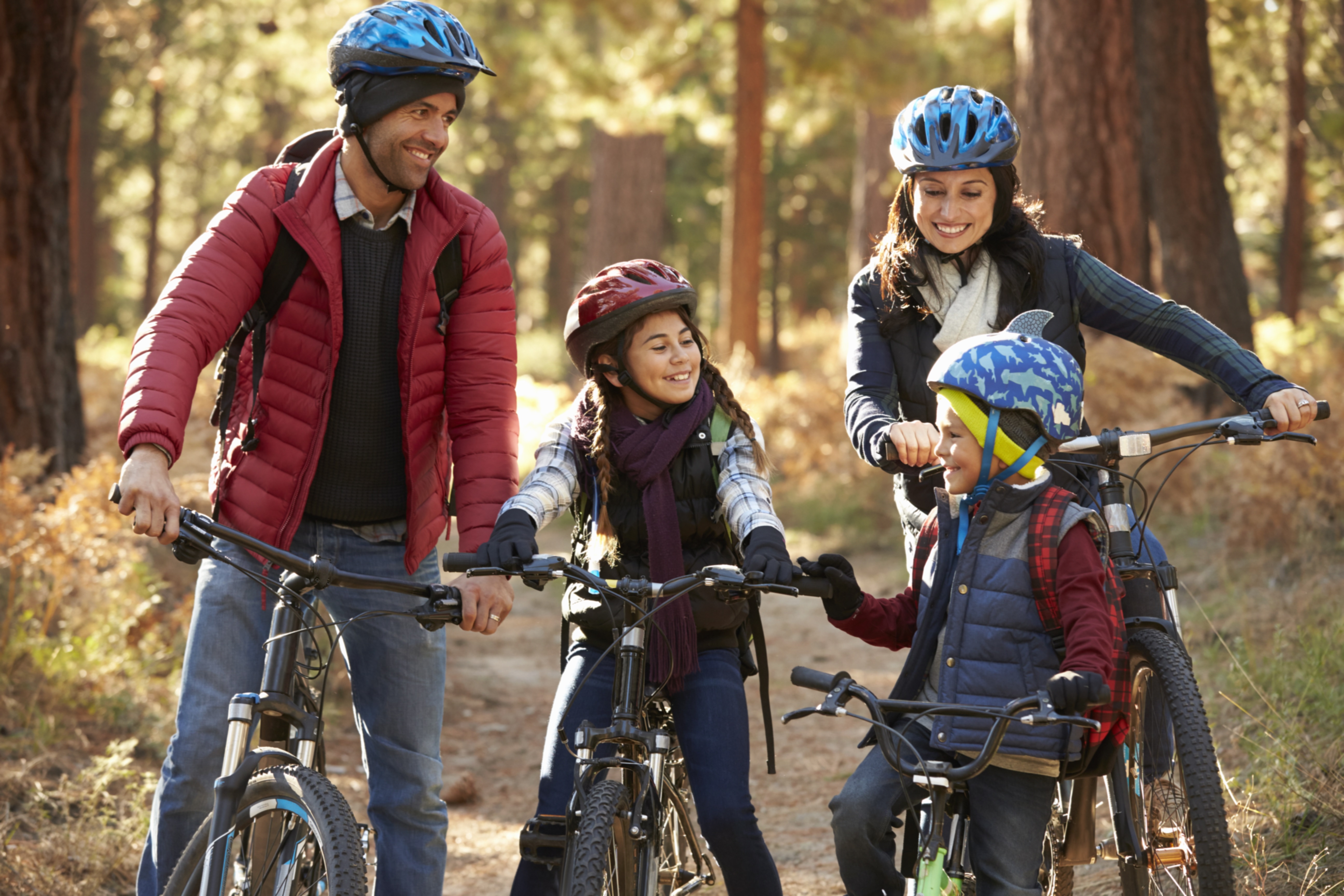 A man, a woman, a boy and a girl sit on bikes, looking toward each other and smiling. Each is wearing a jacket and a helmet. Behind them is a dirt path and several trees.